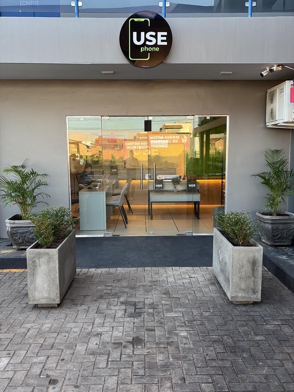 Fachada da USE phone em Boa Vista: porta de vidro, vasos de plantas e o letreiro redondo preto com o logo verde